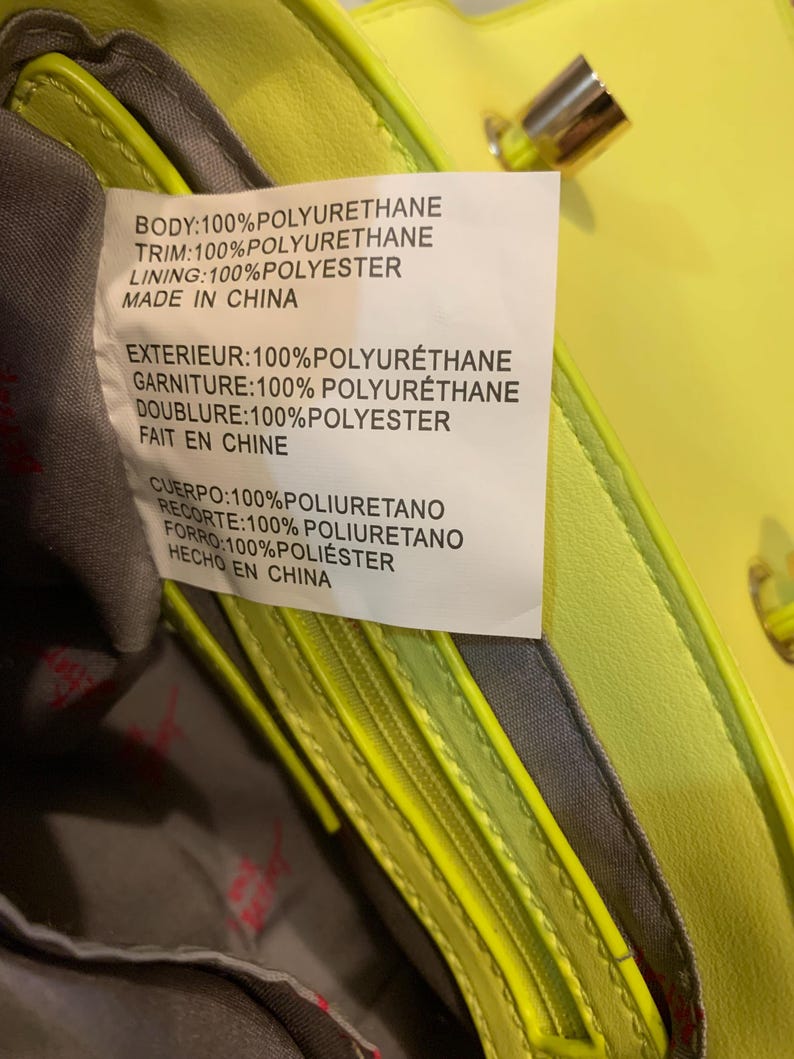 May include: A bright neon yellow handbag with a grey interior lining. A white care label is attached to the inside, displaying material composition in English, French, and Spanish, stating 100% polyurethane body and trim with 100% polyester lining.