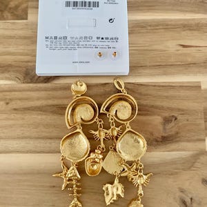 Puede incluir: Pendientes dorados con motivos marinos. Cuentan con m&uacute;ltiples colgantes, incluyendo esqueletos de peces, estrellas de mar y formas de hojas. Los pendientes cuelgan frente a una etiqueta blanca con el nombre de la marca e informaci&oacute;n de tallas.