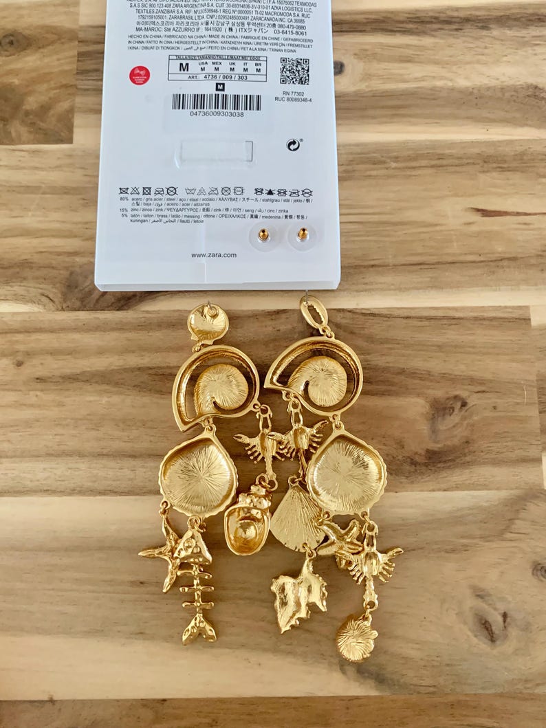Puede incluir: Pendientes dorados con motivos marinos. Cuentan con m&uacute;ltiples colgantes, incluyendo esqueletos de peces, estrellas de mar y formas de hojas. Los pendientes cuelgan frente a una etiqueta blanca con el nombre de la marca e informaci&oacute;n de tallas.