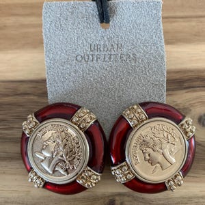 May include: A pair of red and gold-toned clip-on earrings. Each earring features a coin-like design with a profile and laurel wreath, surrounded by a red border and gold accents. The tag behind the earrings reads "URBAN OUTFITTERS".