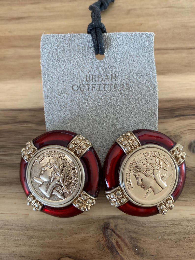 May include: A pair of red and gold-toned clip-on earrings. Each earring features a coin-like design with a profile and laurel wreath, surrounded by a red border and gold accents. The tag behind the earrings reads "URBAN OUTFITTERS".