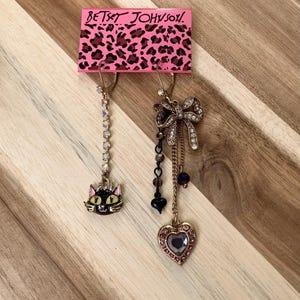 Può includere: Un paio di orecchini Betsey Johnson. Un orecchino presenta un ciondolo a forma di gatto nero con occhi gialli e una catena tempestata di cristalli. L'altro orecchino presenta un fiocco dorato con cristalli, un ciondolo a forma di cuore con un cristallo viola e una catena con perline nere e blu.