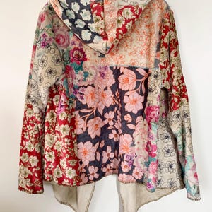 May include: A hooded jacket with a patchwork design featuring various floral patterns in shades of red, pink, and cream. The jacket has a beige lining and a unique, open front design. The hood is also patterned with flowers.