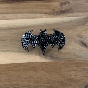 Op de afbeelding: Een zwarte metalen broche in de vorm van een vleermuis, versierd met zwarte strass.