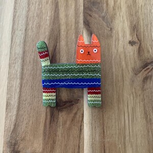 May include: A colorful cat-shaped brooch with an orange head, green and blue body, and striped legs and tail. The brooch has a glittery finish and is made of acrylic. The cat is standing upright.