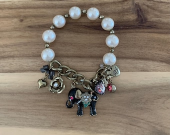 May include: A pearl bracelet with gold-tone chain and charms. The charms include a black elephant with floral accents, a heart, a rose, and a small bow. The bracelet is on a wooden surface.