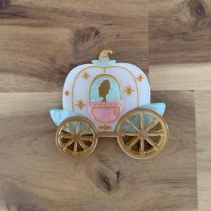 May include: A light-coloured carriage brooch with gold wheels and accents. The carriage features a silhouette of a person inside, with star details and light blue accents. The brooch is set against a wooden surface.