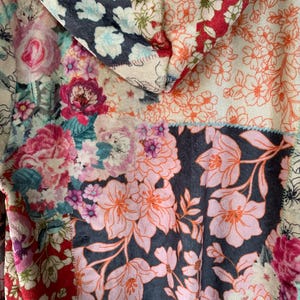 May include: A patchwork hoodie featuring a variety of floral patterns in shades of pink, red, blue, and white. The design includes a hood and is made of a soft, textured fabric. The overall aesthetic is a mix of vintage and bohemian styles.