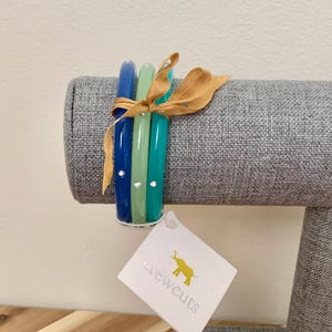 Puede incluir: Tres pulseras de brazalete apiladas en tonos de azul y verde con diamantes de imitación transparentes. Las pulseras están en un expositor gris y tienen una etiqueta blanca con el texto "the crewcuts" en amarillo.