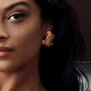 May include: A close-up of a person wearing a red and gold earring. The earring has a circular, ornate design with a gold frame and a red center. The earring is a vintage style.