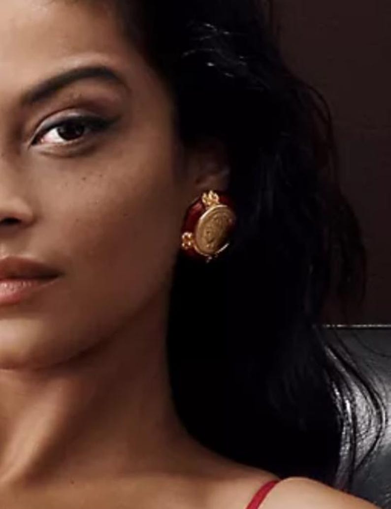 May include: A close-up of a person wearing a red and gold earring. The earring has a circular, ornate design with a gold frame and a red center. The earring is a vintage style.