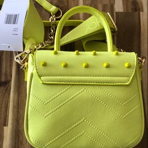May include: A neon yellow handbag featuring a top handle, a detachable shoulder strap with embossed text, and a flap closure adorned with five matching yellow studs. The front of the bag displays a quilted chevron pattern and a white product tag.