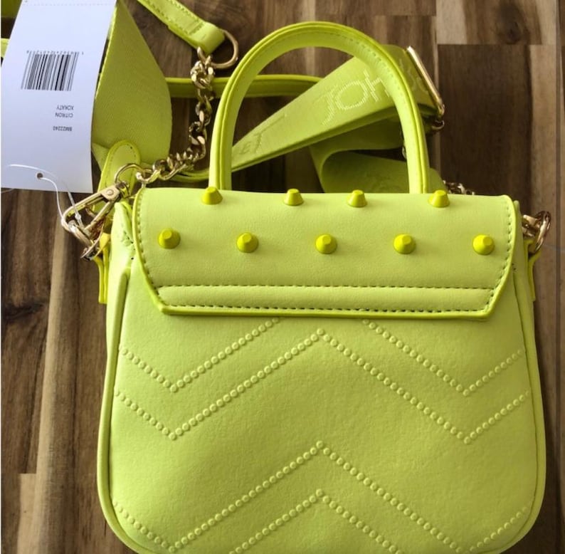 May include: A neon yellow handbag featuring a top handle, a detachable shoulder strap with embossed text, and a flap closure adorned with five matching yellow studs. The front of the bag displays a quilted chevron pattern and a white product tag.