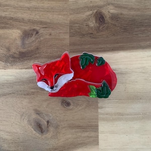 May include: A red fox brooch with white accents on its face and chest, and green leaves. The fox is curled up, appearing to be sleeping. The brooch is made of a shiny material.