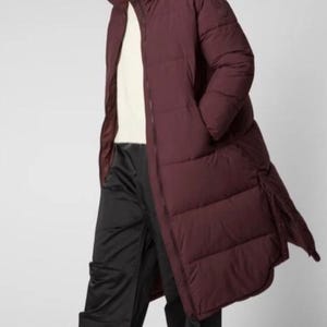 Puede incluir: Abrigo largo acolchado burdeos con capucha grande. El abrigo se lleva sobre un jersey blanco y pantalones negros. El modelo también lleva una gorra negra y zuecos negros con suelas de madera. El abrigo mide unos 140 cm de largo.
