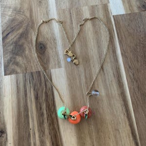 Puede incluir: Un collar de cadena dorada con tres dijes de fruta. Los dijes son verdes, naranjas y rojos y tienen pequeñas hojas verdes adjuntas.