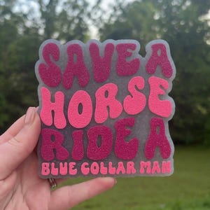 May include: A gray, glittery, square-shaped air freshener with the text "SAVE A HORSE RIDE A BLUE COLLAR MAN" in shades of pink. The air freshener is held in a hand against a blurred green background.