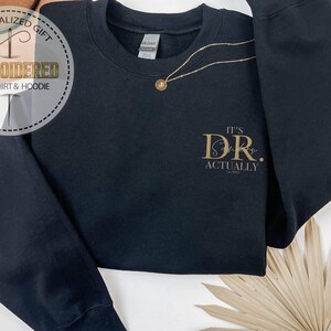 May include: Black sweatshirt with the text "It's Dr. Actually" in gold lettering. A gold necklace with a pendant is draped on the sweatshirt. The sweatshirt has a crew neck and long sleeves. The image also includes a circular logo that says "Personalized Gift Embroidered Sweatshirt & Hoodie."
