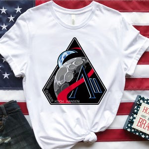 May include: White t-shirt featuring a black, red, and blue graphic of a moon, Earth, and the letter "A". The shirt is displayed on an American flag. A pair of denim shorts and a small book with the text "HOME OF THE BRAVE" are also in the image.