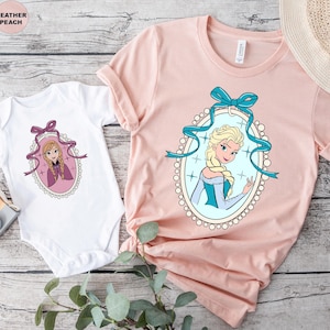 May include: A white baby onesie and a light pink t-shirt featuring cartoon portraits of a princess in an oval frame with a blue bow. The t-shirt has short sleeves and a round neckline. The background is a light wood grain.