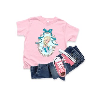 May include: A light pink t-shirt featuring a cartoon image of a blonde woman in a blue dress, framed by a blue ribbon and an oval border. The shirt is paired with blue jeans and pink sneakers.