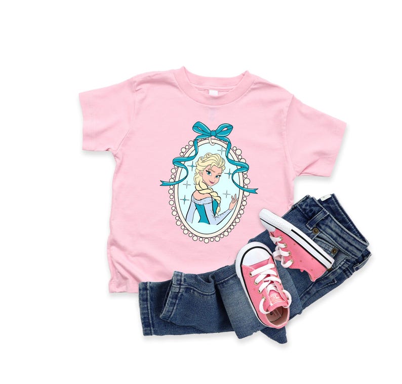 May include: A light pink t-shirt featuring a cartoon image of a blonde woman in a blue dress, framed by a blue ribbon and an oval border. The shirt is paired with blue jeans and pink sneakers.