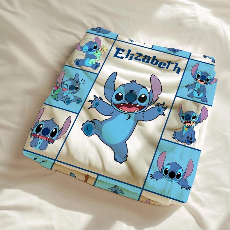 May include: A folded, light-colored blanket featuring a repeating pattern of a blue cartoon character in various poses. The central image shows the character with arms outstretched. The name "Elizabeth" is printed in blue.
