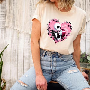 Puede incluir: Una camiseta color crema con un dise&ntilde;o en forma de coraz&oacute;n con Jack Skellington y Sally de The Nightmare Before Christmas. El coraz&oacute;n es rosa y est&aacute; rodeado de rosas rosas, con las palabras "Love You to Death".