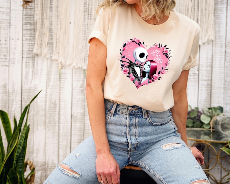 Puede incluir: Una camiseta color crema con un dise&ntilde;o en forma de coraz&oacute;n con Jack Skellington y Sally de The Nightmare Before Christmas. El coraz&oacute;n es rosa y est&aacute; rodeado de rosas rosas, con las palabras "Love You to Death".
