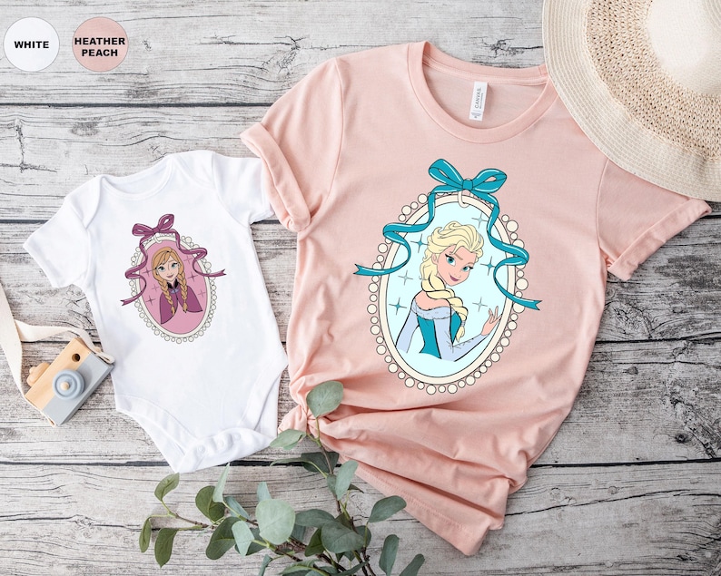 May include: A white baby onesie and a light pink t-shirt featuring cartoon portraits of a princess in an oval frame with a blue bow. The t-shirt has short sleeves and a round neckline. The background is a light wood grain.