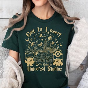 May include: A dark green t-shirt with a gold graphic of the Hogwarts castle from the Harry Potter series. The graphic also includes two Minions from the Despicable Me franchise. The text "Get in Loser! We're going to Universal Studios" is printed on the shirt.