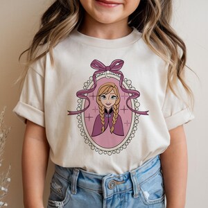 May include: A cream-colored t-shirt featuring a cartoon portrait of a character with blonde braids, wearing a purple cape, inside a pink oval frame with a purple bow and ribbon. The shirt is worn by a child.