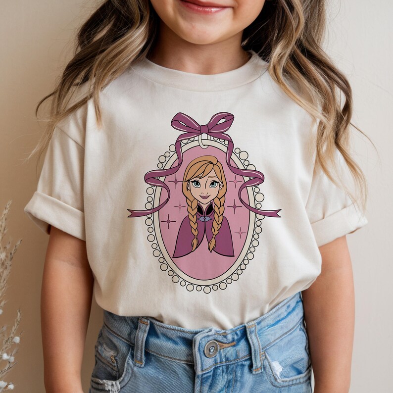 May include: A cream-colored t-shirt featuring a cartoon portrait of a character with blonde braids, wearing a purple cape, inside a pink oval frame with a purple bow and ribbon. The shirt is worn by a child.