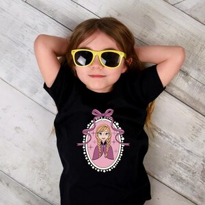 May include: A black t-shirt featuring a cartoon image of a princess in a pink oval frame with a purple bow. The girl in the image is wearing yellow sunglasses and is lying on a wooden surface.