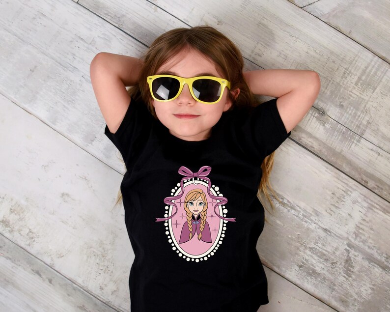 May include: A black t-shirt featuring a cartoon image of a princess in a pink oval frame with a purple bow. The girl in the image is wearing yellow sunglasses and is lying on a wooden surface.