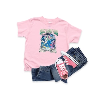 May include: A pink Buzz Lightyear t-shirt with a graphic of Buzz Lightyear and other characters from the Disney Toy Story movie. The shirt is paired with blue denim jeans and pink sneakers.