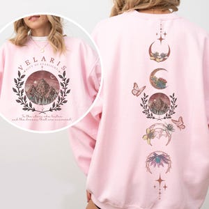 Velaris-themed pink t-shirt with front design of mountains, stars, and 'To the stars who listen' quote. Back highlights moon phases, butterflies, and floral art, blending celestial and fantasy vibes in a stylish, soft fabric.