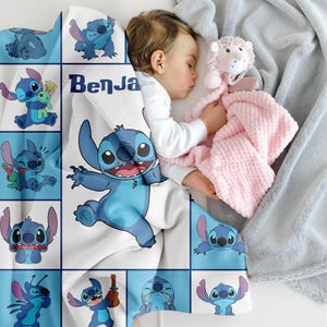May include: A baby sleeps under a white and blue blanket featuring the character Stitch from Lilo & Stitch. The blanket has a grid pattern with various Stitch illustrations and the name "Benja" printed on it. A pink plush toy and a gray blanket are also visible.