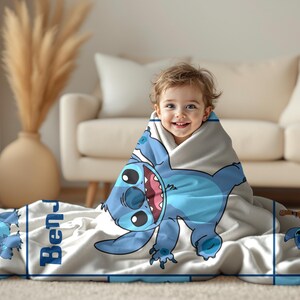 May include: A light gray blanket featuring a large blue cartoon character with big eyes and a wide grin. The blanket has a blue border and the name "Ben" printed on it. A child is wrapped in the blanket.