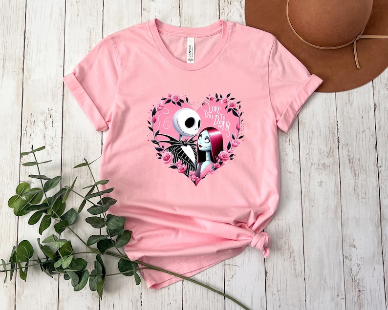 Puede incluir: Camiseta rosa claro con un dise&ntilde;o de Jack Skellington y Sally de Pesadilla antes de Navidad dentro de una corona floral en forma de coraz&oacute;n. El texto "Love You to Death" est&aacute; sobre los personajes. La camiseta es de material suave.