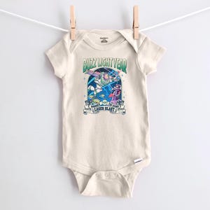 May include: A cream-colored baby onesie with a graphic featuring Buzz Lightyear and other characters from Toy Story. The text "Buzz Lightyear" and "Disney Pixar Toy Story Laser Blast" is printed on the front.