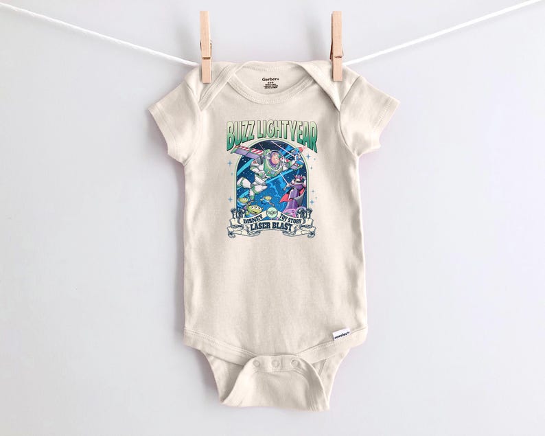 May include: A cream-colored baby onesie with a graphic featuring Buzz Lightyear and other characters from Toy Story. The text "Buzz Lightyear" and "Disney Pixar Toy Story Laser Blast" is printed on the front.