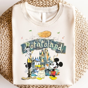 May include: A beige t-shirt with a cartoon illustration of Mickey Mouse, Donald Duck, and Goofy in front of a castle with the text "Potato Land".