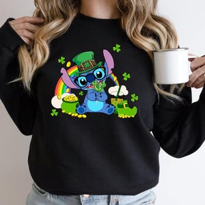 May include: Black sweatshirt featuring a cartoon Stitch character dressed for St. Patrick's Day. Stitch is wearing a green hat and holding a shamrock. A rainbow, pot of gold, and green shamrocks are also featured.