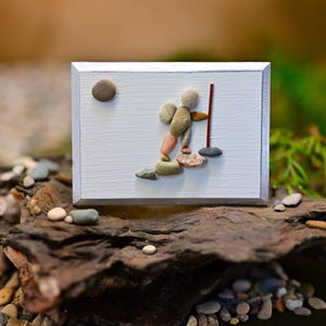 May include: A rectangular white wooden plaque featuring a pebble art composition of a figure hiking with a backpack and a walking stick. A small round brown pebble represents the sun in the upper left corner of the white painted surface.