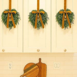 May include: Three small green evergreen wreaths hanging from off-white kitchen cabinets by long mustard-yellow velvet ribbons. Each wreath features a central mustard-yellow bow and a small metallic bell. Two wooden cutting boards sit on the counter.