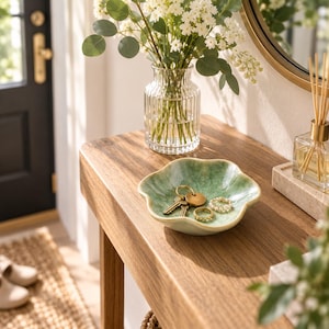 May include: A small, scalloped, light green ceramic bowl with a crackle glaze finish. The bowl contains a set of metal keys on a gold ring and two small, beaded rings. The bowl is positioned on a wooden surface near a glass vase of flowers.
