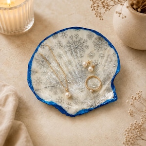 May include: A decorative shell-shaped trinket dish featuring a white surface with a silver snowflake pattern and a vibrant blue painted edge. The dish holds a gold chain necklace with a pearl pendant, a gold ring with a pearl, and two pearl earrings.