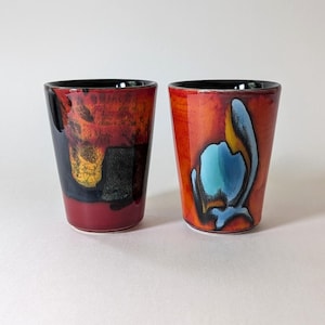 May include: Two ceramic tumblers with a glossy finish. The left tumbler features a blend of red, black, and yellow abstract patches. The right tumbler displays a vibrant orange background with a prominent blue and black abstract shape on the front.