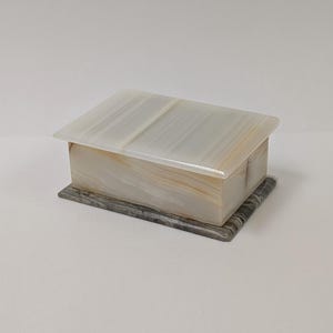 May include: A rectangular stone box with a smooth, translucent, off-white lid featuring subtle linear banding. The main body of the box displays soft beige and cream veining, while the base is composed of a darker, grey-toned stone with distinct patterns.